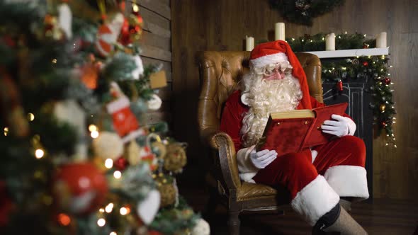 Santa Claus Pulls Out Old Red Silk Album Near Christmas Tree with Toys and Lights