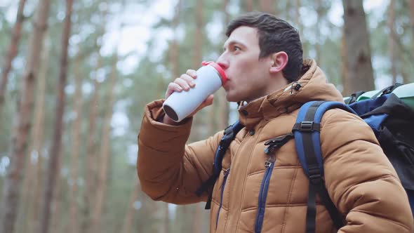 Young man traveler drinking hot coffee or tea in thermos mug in forest