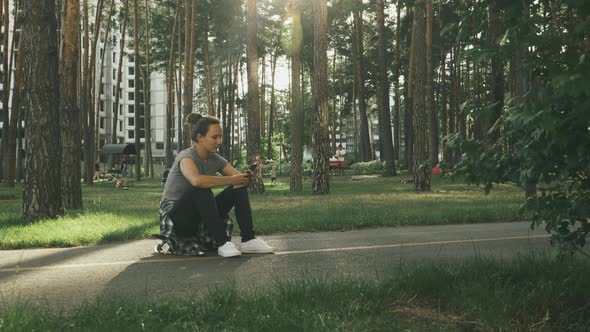 Happy hipster woman in jeans and plaid shirt with skateboard and phone sitting in park at sunset.