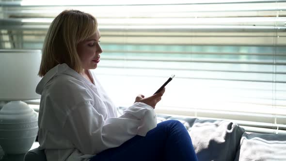 a Young Woman Uses a Smartphone and Slowly Lies on the Sofa in the Living Room at Home