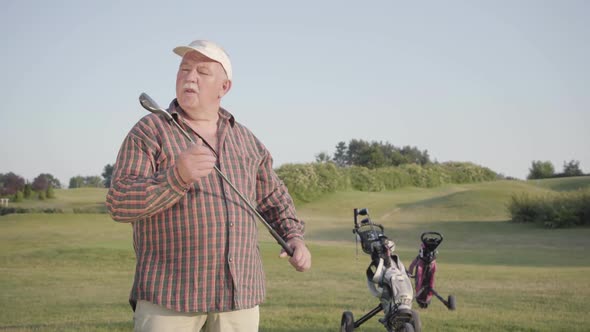 Senior Man Blows Dust Off a Golf Club, Rubs It Before a Game. Old Gentleman Playing Game Outdoors