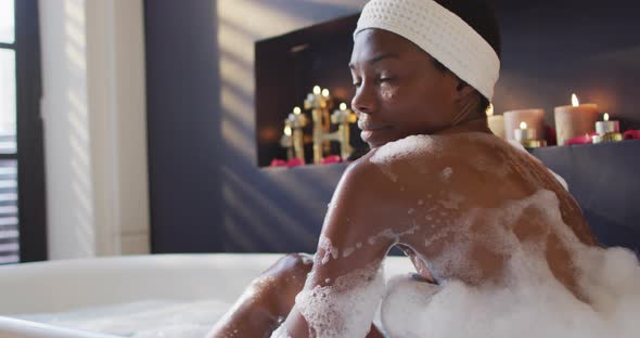 Smiling african american woman with towel taking bath and touching her skin in bathroom