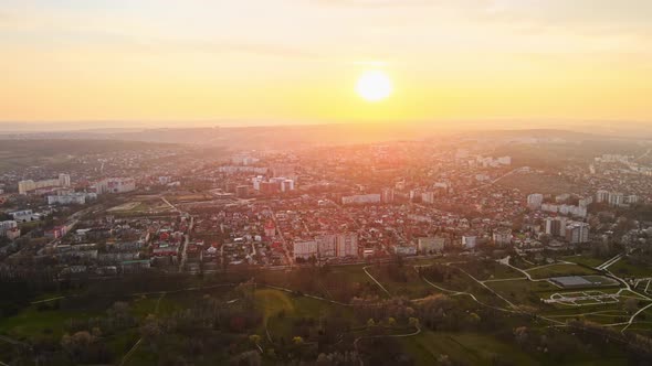 Aerial drone view of Chisinau at sunset. Multiple buildings, trees, park with a lake, road with movi