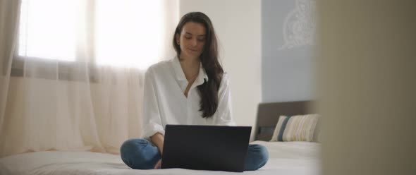 A young woman working on her laptop, while sitting in bed.