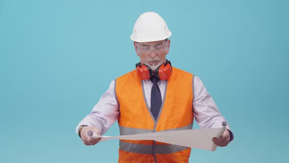 Engineer in glasses, vest, helmet and headphones unwraps a bundle of drawings and studies the scheme