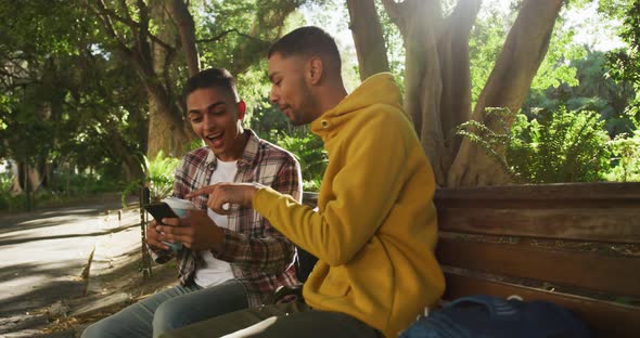 Two happy mixed race male friends sitting and using smartphone in park with backpacks