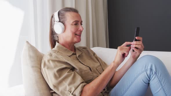 Caucasian senior woman wearing headphones using smartphone sitting on the couch at home