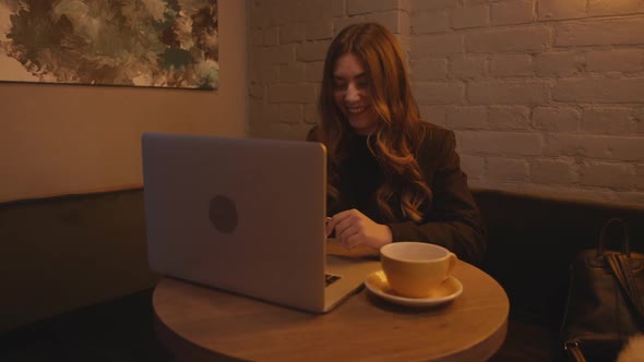 Elegant Young Girl in Black Clothes Communicates in a Laptop and Laughs While Sitting on a Sofa in a
