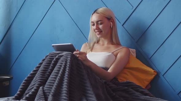 Girl with Headphones is Sitting on Bed Covered with Blanket and Talking Video Link on Electronic