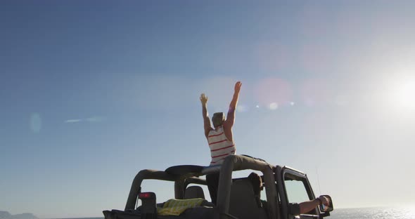 Happy caucasian gay male couple in car raising arms and waving on sunny day at the beach