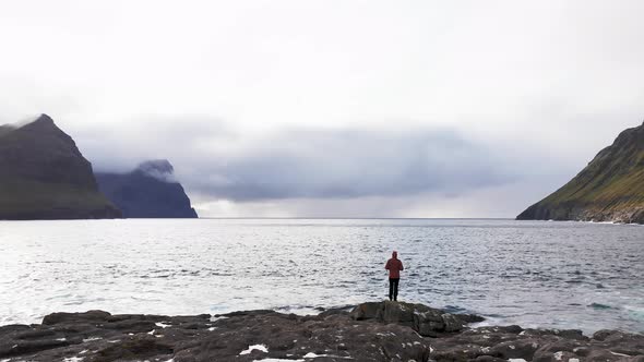 Aerial View of Unrecognizable Woman Stands on Rock Cliff Looking at Wilderness Background of Faroe