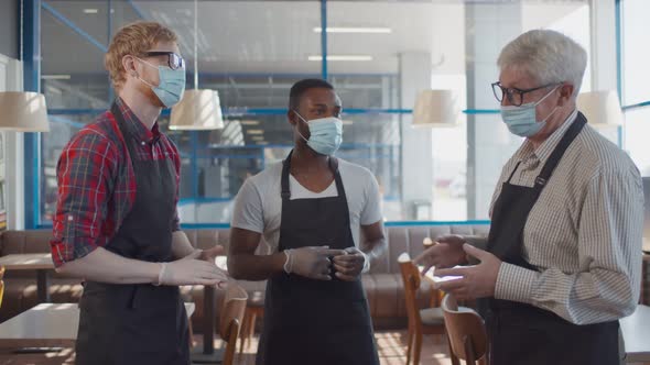 Seinor Cafe Owner Instructing Young Diverse Waiters in Safety Mask and Gloves