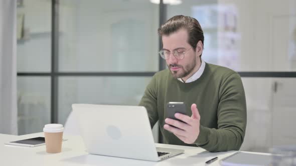 Young Businessman Using Smartphone While Using Laptop in Office