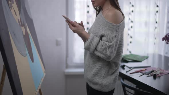 Unrecognizable Young Woman Standing at Easel with Picture Surfing Social Media on Smartphone