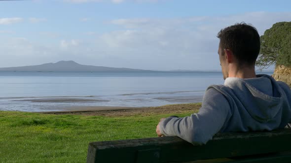 Relaxed young caucasian man drinking coffee sitting on a bench at the beach  looking at Rangitoto is