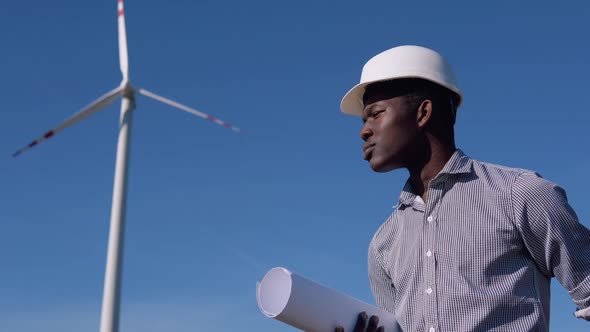 African American Electrical Engineer in a Helmet and with a Drawing in His Hand Stands on the