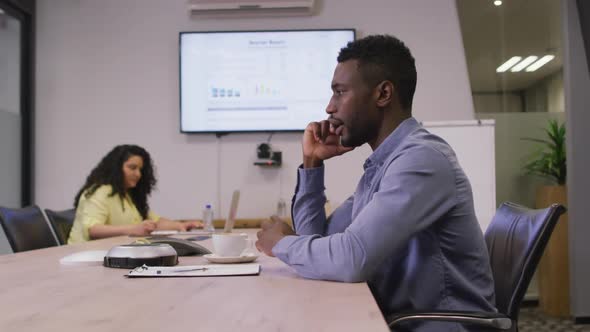 African american businessman sitting at desk, talking on smartphone in modern office