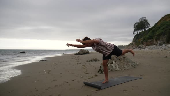 A young man yoga instructor doing difficult balancing poses on the beach with ocean waves in slow mo