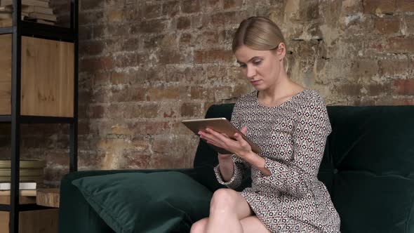 Woman scrolling on tablet at home, uses technology. Woman sits on sofa, she's using tablet computer.