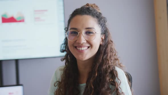 Portrait of smiling biracial businesswoman wearing glasses in meeting room with screen in background