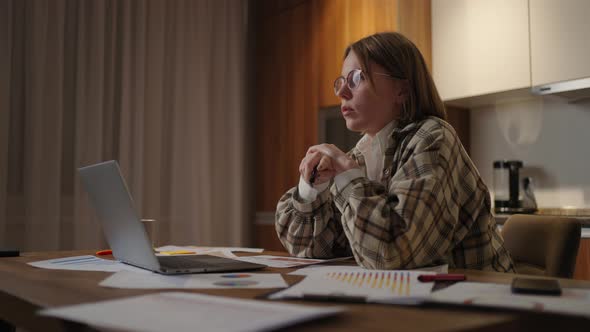 A Focused Woman with Glasses Working on Financial Documents Sitting in the Workplace Using a Laptop