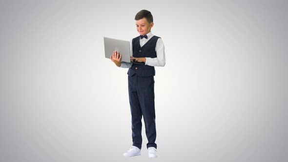 Smiling Boy in a Bow Tie and Waistcoat Using Laptop Computer While Standing on Gradient Background