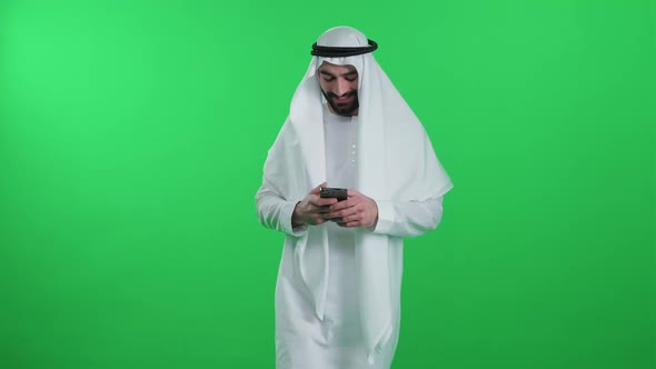 Arab Man in a White Kandura on the Green Background Walks Using a Smartphone and Surf the Internet