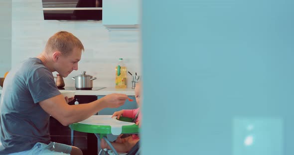 Loving Young Dad Holding a Spoon and Feeding the Baby in the Kitchen