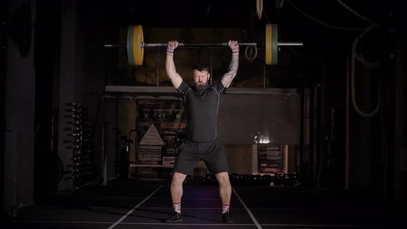 Muscular Man with Beard Does Overhead Deadlift with Barbell on Dark Background