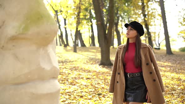 Fashion Girl in Stylish Clothes Posing at Sculpture in Sunny Autumn Park