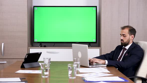 Businessman in Meeting Room Next To a Green Screen TV