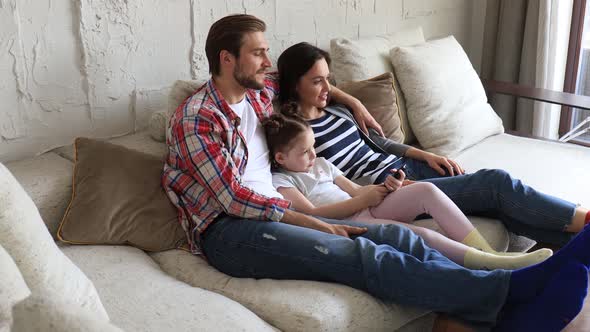 Beautiful happy young parents and their little daughter watching TV and smiling while sitting on sof