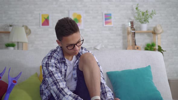 Young Asian Man Sitting on the Couch Puts a Tightening Elastic Bandage on a Sick Knee Close Up
