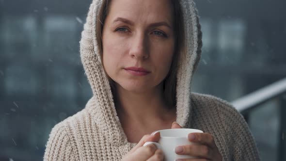 Caucasian Woman Stays on Balcony During Snowfall with Cup of Hot Coffee or Tea