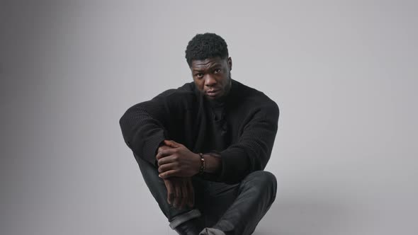 Serious Young African American Man Looking Up to the Camera Sitting on the Floor in Studio Grey