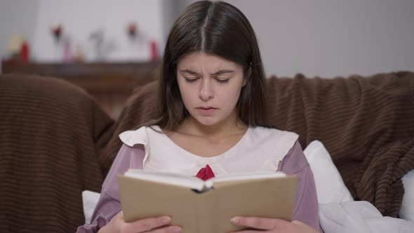 Portrait of Concentrated Young Female Student Reading Sitting on Couch with Blurred Unrecognizable