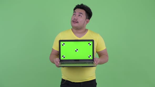 Happy Young Overweight Asian Man Thinking While Showing Laptop