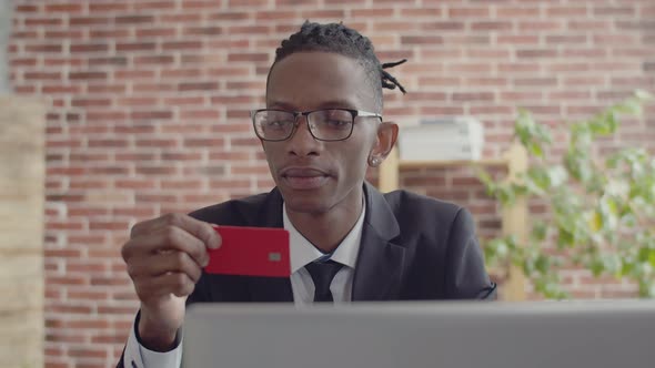 Black man businessman in glasses and a suit with a tie holds red bank card in hand