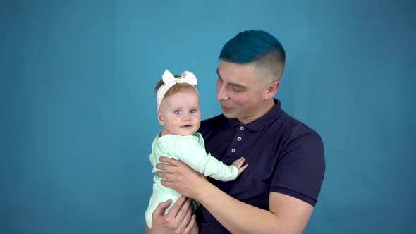 A Young Father with Blue Hair Holds a Baby in His Arms. Alternative Man with a Child Look at the