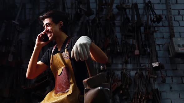 Authentic Blacksmith Man Wear Leather Apron in Dark Indoors Studio Talking By Phone and Laughing