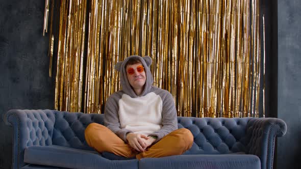 Young Man in Hood with Ears and Bright Glasses Sitting on Sofa Against Wall with Golden Tinsel