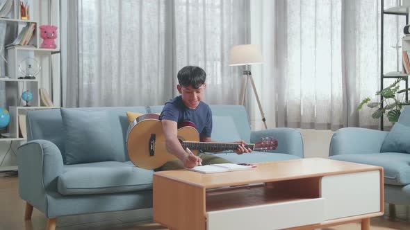 Asian Boy Composer With A Guitar Thinking Before Composing Music On Paper At Home