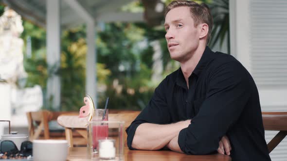 Upset Man Sits in Stylish Cafe with Freshly Squeezed Juice and Looks at Camera