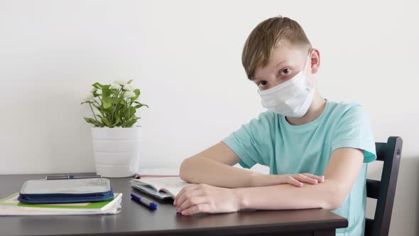 A Young Boy in a Face Mask Looks Up From a School Notebook To the Camera and Points at It at a Table