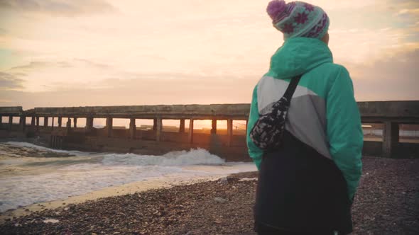 Attractive Young Woman in a Warm Jacket Walking on the Stone Beach at Sunset