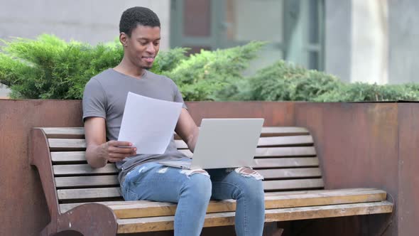 Young African Man Working on Papers and Laptop on Bench