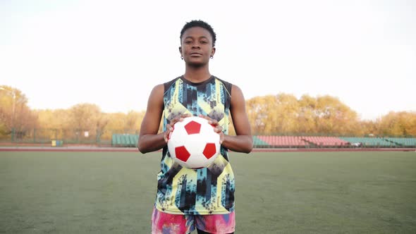 A Serious Young Black Girl in a Sleeveless Tshirt Stands on a Soccer Field and Holds a Ball in Her