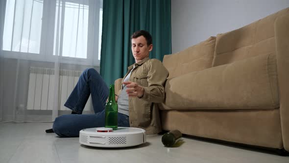 White Robot Vacuum Cleaner Delivers Alcohol Beer Bottle to Brunet Man Sitting on Floor Near Sofa in