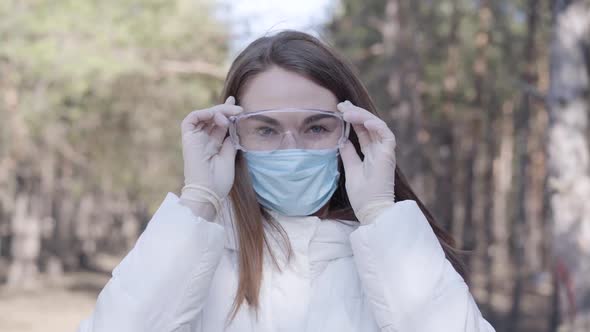 Serious Caucasian Girl in Protective Gloves and Face Mask Putting on Transparent Eyeglasses