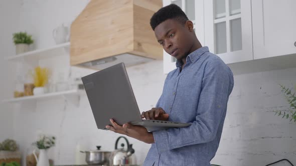 Middle Shot of Concentrated Young Man Standing in Kitchen Typing on Laptop Keyboard